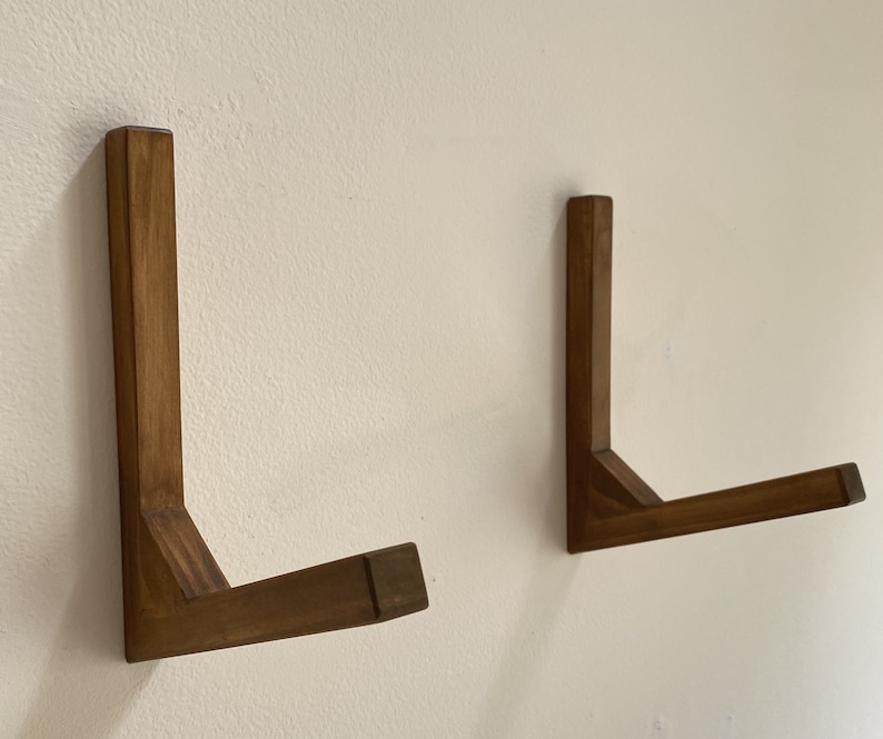 May include: Two wooden wall mounted L-shaped shelves. The shelves are made of dark brown wood and are mounted on a white wall.