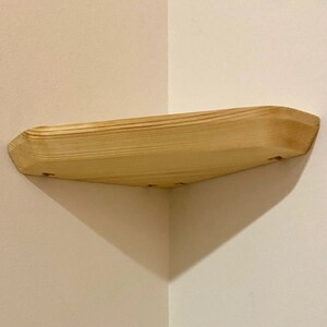 May include: A natural wood corner shelf with a rounded edge and a light wood finish. The shelf is mounted to a wall with screws.