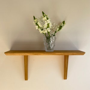 May include: A wooden shelf with two angled supports, featuring a clear glass vase with white flowers.