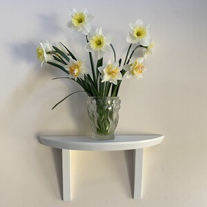 Puede incluir: Un jarrón de cristal transparente lleno de narcisos blancos y amarillos. El jarrón está sobre una estantería blanca semicircular fijada a una pared clara. Los narcisos tienen tallos y hojas verdes.