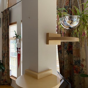 May include: A light wood, wall-mounted shelf and a round, light wood shelf attached to a white column. A hanging, mirrored glass planter with a plant is suspended from the ceiling. The room has a window with blinds and floral curtains.