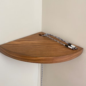 May include: A wooden corner shelf with a rounded edge and a hole for cords. Two gray and black cords are threaded through the hole.