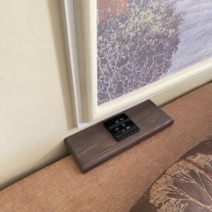 以下が含まれることがあります： 複数のUSBポートとコンセントを備えた黒い電源タップが付いた、濃い茶色の木製棚。 棚は長方形で、白い縁のある額入りの絵の近くのベージュ色の表面に置かれています。