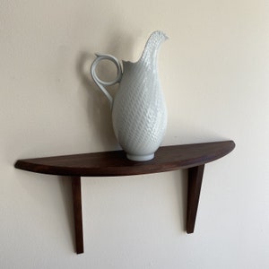 Puede incluir: Una jarra de cerámica blanca con una superficie texturizada y un asa curva se encuentra sobre una estantería de madera semicircular de color marrón oscuro. La estantería está montada en una pared color crema. La jarra tiene un pico alargado único.