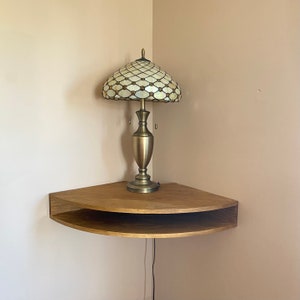 May include: A wooden corner shelf with two tiers, featuring a decorative lamp with a stained glass shade. The lamp has a gold base and a white and gold patterned shade.