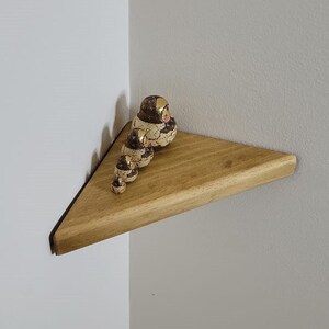 Op de afbeelding: Een houten hoekplank met een set van in elkaar passende poppen in tinten bruin en goud. De poppen zijn in een rij gerangschikt, met de grootste pop bovenaan en de kleinste pop onderaan.