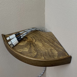 以下が含まれることがあります： 濃い茶色のステインを施した木製のコーナーシェルフ。白い編み込みの充電ケーブルが3本、シェルフのコンセントに差し込まれています。