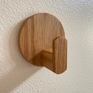 Puede incluir: Un gancho de pared de madera con una base redonda y un solo gancho que se extiende hacia afuera. El gancho está hecho de madera marrón claro con un patrón de veta natural.