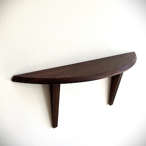 Può includere: Una mensola in legno marrone scuro, a forma di semicerchio, montata su una parete bianca. La mensola ha un bordo anteriore liscio e curvo ed è sostenuta da due gambe angolate. La venatura del legno è visibile, aggiungendo consistenza al design.