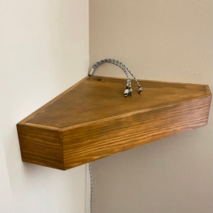Puede incluir: Una estantería de esquina de madera con un tinte marrón oscuro. La estantería tiene forma triangular y está montada en una pared. Tres cables negros y blancos cuelgan de la estantería.