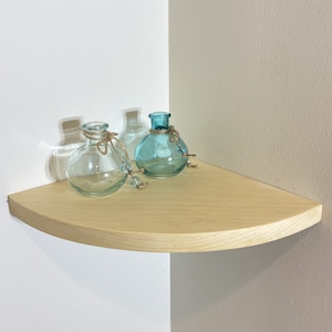 May include: A light wood corner shelf with a rounded edge, featuring three glass vases. Two of the vases are a light blue color, and one is clear. The vases are decorated with twine.