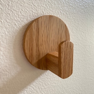 Puede incluir: Un gancho de pared de madera con una base redonda y un solo gancho. El gancho está hecho de madera marrón claro con un patrón de veta natural.