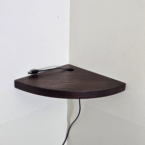 May include: A dark brown, triangular corner shelf made of wood. A black cable with a USB connector is plugged into the shelf and runs down the wall. The shelf has a hole for the cable.