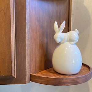May include: White ceramic bunny figurine sitting on a round white ceramic base. The bunny is in a curled up position with its head resting on the base. The figurine is on a wooden shelf.