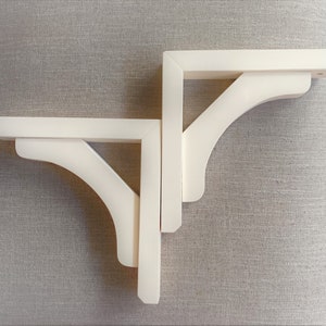 以下が含まれることがあります： 2つのオフホワイトのL字型棚受け。各ブラケットには、湾曲したサポートピースと平らな上部と側面があります。ブラケットは木製で、棚を支えるために壁に取り付けるように設計されています。