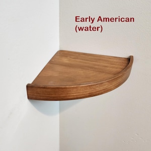 Puede incluir: Un estante de esquina de madera con un borde redondeado. El estante está teñido de un color marrón oscuro y está etiquetado como "Early American (water)".