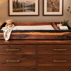 May include: A rectangular wooden changing tray with a dark live edge border, featuring a white textured changing pad, a small teddy bear, and a side compartment containing white cloths and a small bottle, resting on a six-drawer wooden dresser.