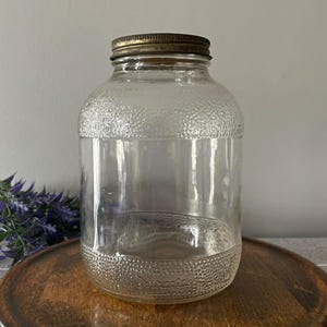 Peut inclure: Un bocal en verre transparent avec une surface texturée et un couvercle en métal. Le bocal a un corps large et un col étroit. Le couvercle est de couleur or terni. Le bocal est posé sur un plateau en bois.