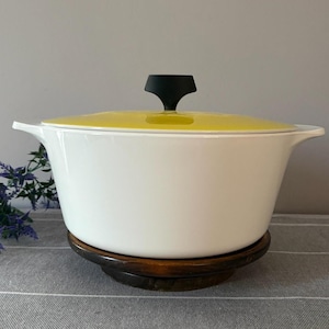 May include: A white enameled cast iron Dutch oven with a bright yellow lid and a black handle. The pot sits on a dark wooden trivet. The cookware is designed for cooking and serving.