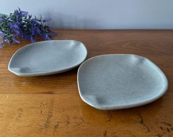 May include: Two gray ceramic plates with a slightly irregular, organic shape. The plates are a muted gray color and have a textured surface. A sprig of purple flowers is in the background. The plates are on a wooden surface.