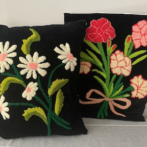 May include: Two black velvet throw pillows with embroidered floral designs. One pillow features a bouquet of pink and red carnations, while the other features a cluster of white daisies.