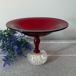 May include: A deep red glass pedestal bowl with a clear, textured base. The bowl has a wide, shallow design and a dark rim. The stem is a deep red colour. The base is clear glass with a bubble design.