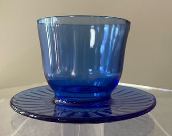 May include: A cobalt blue glass cup and saucer set. The cup has a rounded shape and the saucer has a fluted edge.