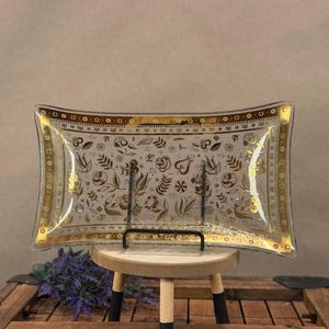 May include: A rectangular glass tray with curved edges featuring a gold-toned border and a central pattern of gold floral and leaf motifs. The clear glass surface displays a repeating design of various botanical elements set against a transparent background.