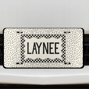 May include: A personalized license plate with the name "LAYNEE" in bold black letters. The plate has a cream background with black polka dots and a checkered border. The plate is mounted on a white car.
