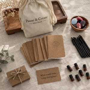 May include: A collection of self-care items, including a drawstring bag with "Pause & Create - Mini Reset" text, small notebooks, essential oil bottles, pens, and a wooden bowl with stones. A card reads "What would make today feel lighter?"