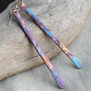 May include: A pair of handcrafted, long, rectangular earrings with a hammered texture. The earrings display a vibrant color palette of blues, purples, and oranges. The ear wires are silver. The earrings are displayed on a piece of driftwood.