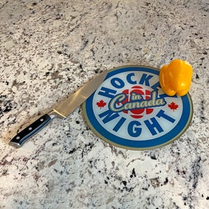 Puede incluir: Una tabla de cortar redonda con un diseño azul y blanco que dice "Hockey Night in Canada". Un pimiento amarillo se encuentra en la tabla junto a un cuchillo de cocina plateado.