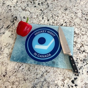 Puede incluir: Tabla de cortar rectangular de vidrio con un diseño de hockey azul y blanco, con la inscripción "HOCKEY NIGHT IN CANADA". Un pimiento rojo y un cuchillo de acero inoxidable con mango negro están sobre la tabla.
