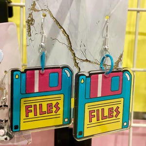 Puede incluir: Un par de pendientes de estilo retro con un diseño de disquete en turquesa, rosa y amarillo. La palabra "FILES" está impresa en la etiqueta amarilla. Los pendientes tienen ganchos plateados y anillos azules.