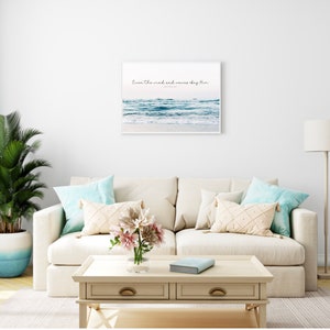 May include: A white sofa with blue and cream cushions in a living room setting. A framed print of a beach scene with the text "Love the wind and waves they bring" hangs on the wall above the sofa. A potted plant and a floor lamp are also visible in the room.