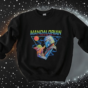 May include: Black sweatshirt featuring a retro-style graphic of the Mandalorian and Grogu. The graphic depicts a triangle with the text "Mandalorian - This is the Way -" and a blue and green image of the Mandalorian holding Grogu.