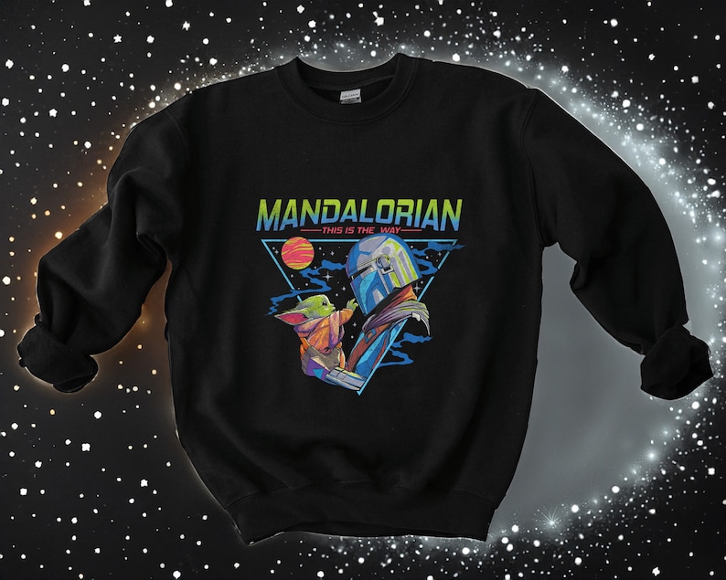 May include: Black sweatshirt featuring a retro-style graphic of the Mandalorian and Grogu. The graphic depicts a triangle with the text "Mandalorian - This is the Way -" and a blue and green image of the Mandalorian holding Grogu.