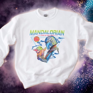 May include: White sweatshirt featuring a graphic print of a Mandalorian character holding a small green alien. The text "MANDALORIAN -THIS IS THE WAY- " is printed above the graphic.
