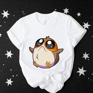 May include: White t-shirt featuring a cartoon image of a brown and orange bird-like creature with large eyes. The creature is wearing a purple headband.