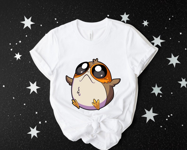 May include: White t-shirt featuring a cartoon image of a brown and orange bird-like creature with large eyes. The creature is wearing a purple headband.