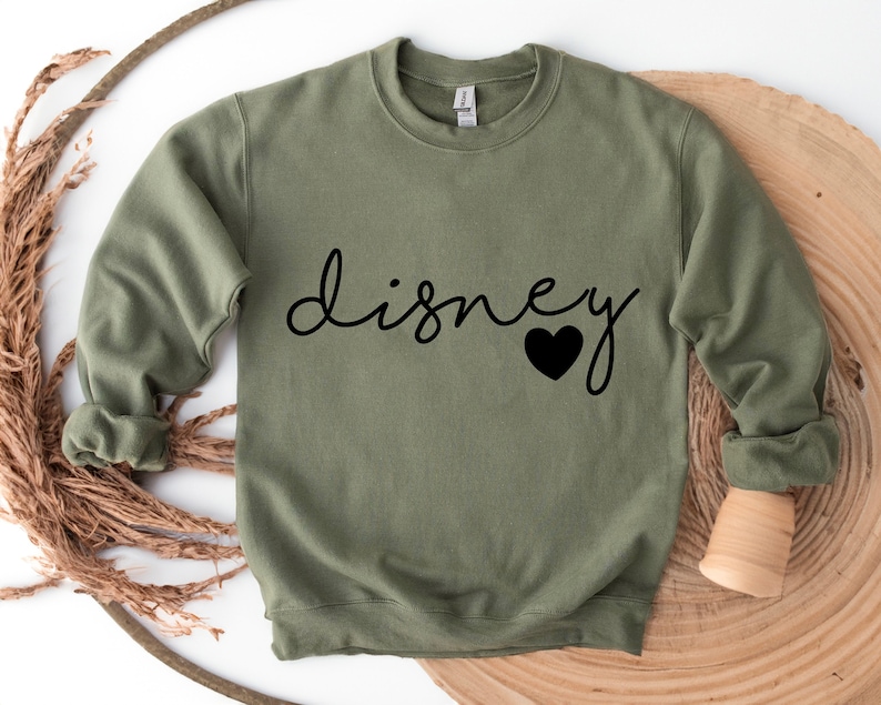 Puede incluir: Una sudadera verde oliva con la palabra "disney" en cursiva y un coraz&oacute;n negro. La parte superior de manga larga con cuello redondo est&aacute; hecha de un material suave. La sudadera se muestra sobre una superficie de madera.