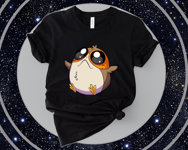 May include: Black t-shirt featuring a cartoon image of a round, white and orange character with large eyes. The character is from the Star Wars franchise.