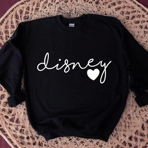 Puede incluir: Sudadera negra con la palabra "disney" en escritura blanca, un coraz&oacute;n blanco reemplaza la letra "o". La sudadera se muestra sobre una superficie beige tejida.
