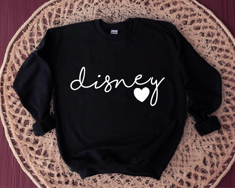 Puede incluir: Sudadera negra con la palabra "disney" en escritura blanca, un coraz&oacute;n blanco reemplaza la letra "o". La sudadera se muestra sobre una superficie beige tejida.