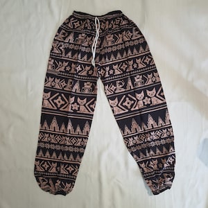 May include: Black and tan patterned pants with a drawstring waist. The pants feature a repeating pattern of stylized cats, buildings, and geometric shapes. The pants have elastic cuffs at the ankles.