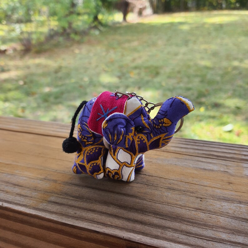 May include: A purple and gold fabric elephant keychain with a pink and blue patterned saddle. The elephant has a black tassel tail.
