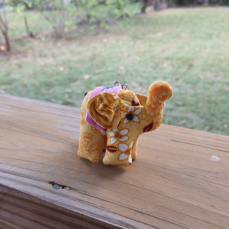 May include: A small yellow and pink fabric elephant with white and gold floral patterns. The elephant has black eyes and a pink saddle.