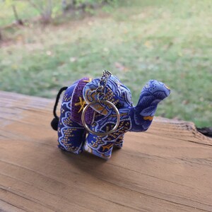 May include: A purple and blue fabric elephant keychain with gold accents and a silver chain. The elephant has a raised trunk and is standing on a wooden surface.