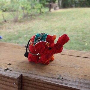 May include: A red fabric elephant keychain with green and yellow accents. The elephant has a small black tassel on its tail.