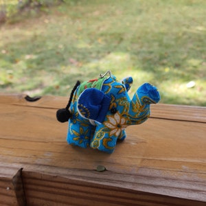 May include: A blue and gold patterned elephant fabric ornament with a black tassel tail. The elephant has a blue ear and a white flower on its trunk.